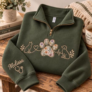 May include: An olive green quarter-zip sweatshirt featuring an embroidered design of a cat, a dog, and a floral-patterned paw print connected by a heartbeat line. The sleeve is embroidered with the name Melissa and a stethoscope icon.