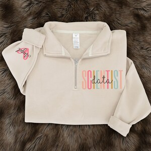 May include: A beige quarter-zip sweatshirt with the word "SCIENTIST" in colorful letters and the word "data" in a script font. The left sleeve has the word "Angel" in pink script with a pink heart.