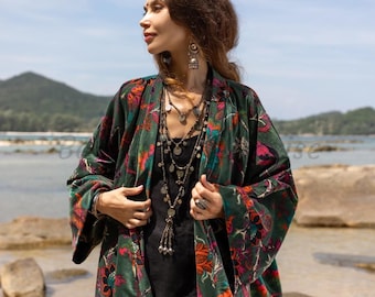 May include: A woman wearing a green velvet kimono with a floral print. The kimono has long sleeves and a wide, open front. She is wearing a black top and jeans underneath. The kimono is decorated with silver jewelry, including a long necklace and earrings.