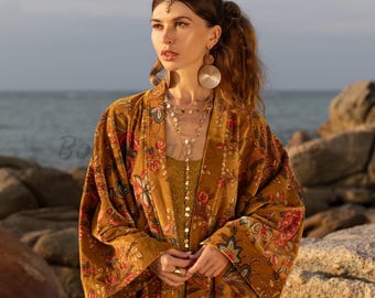 Puede incluir: Kimono con estampado floral color mostaza con mangas anchas. El kimono presenta un patrón vibrante de flores rojas, rosas y azules sobre un fondo dorado. La modelo lleva joyas doradas, incluyendo pendientes, collares y un adorno para la cabeza. El fondo muestra rocas y el mar.