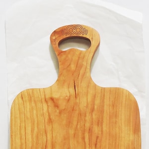 May include: A wooden cutting board with a handle. The board is rectangular with rounded corners and a handle with a decorative design. The wood grain is visible, and the board has a warm, natural color.
