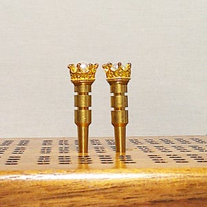 Peut inclure: Deux pions en métal de couleur dorée avec une couronne et une perle blanche sur le dessus. Les pions sont debout sur une surface en bois avec un motif en grille.