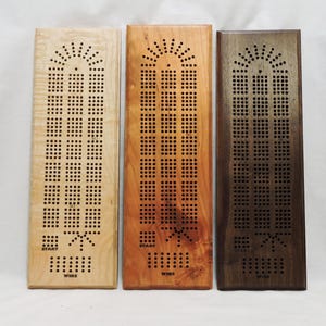 May include: Three wooden cribbage boards in varying shades of wood: light, medium, and dark. Each board has a series of drilled holes for pegging, a starburst design at the top, and the words "START" and "WINS" at the bottom.