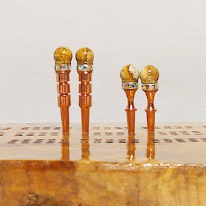 May include: Four decorative wooden dowels with a copper finish and a gemstone top. The gemstones are a light brown color with a speckled pattern and are surrounded by a band of iridescent rhinestones.