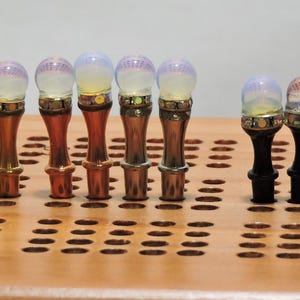 May include: A collection of decorative bottle stoppers with a variety of designs. The stoppers have a bulbous top with a pearlescent or patterned appearance, and a metallic base. The bases are in shades of gold, copper, and black. They are displayed on a wooden cribbage board.