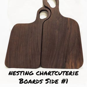 May include: Two nesting charcuterie boards made of dark wood. The boards have a handle with a circular opening. The text "NESTING CHARTCUTERIE BOARDS SIDE #1" is written in black at the bottom of the image.