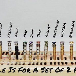 May include: A set of twelve cribbage pegs, each with a different colored gem on top, arranged in a row. The months of the year are written above each peg. The text "SALE IS FOR A SET OF 2 PEGS" is written at the bottom.