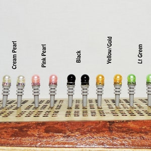 May include: A collection of cribbage pegs in various colors, including cream pearl, pink pearl, black, yellow/gold, and light green. The pegs are displayed on a wooden cribbage board. The text above the pegs indicates the color of each peg.
