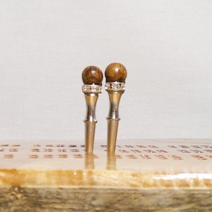May include: Two cribbage pegs with wooden ball tops and silver-colored shafts. The pegs are inserted into a wooden cribbage board with a light-colored surface and small holes. The pegs are a decorative accessory for the classic board game.