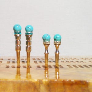 May include: Four turquoise and gold decorative tacks with a round top. The tacks are on a wooden surface with a grid pattern.