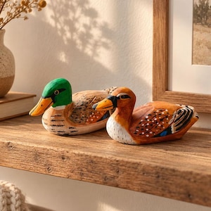 Könnte beinhalten: Zwei handbemalte Entenfiguren aus Holz. Die linke Ente hat einen leuchtend grünen Kopf, einen gelben Schnabel und einen braun gemusterten Körper. Die rechte Ente hat einen braunen Kopf, einen orangefarbenen Schnabel und weiß-schwarz gepunktete Details.