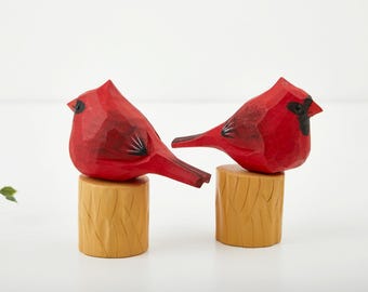 Puede incluir: Dos figuras de pájaros cardenales de madera tallada. Los pájaros son de color rojo brillante con detalles en negro y se asientan sobre pequeños tocones de madera de color marrón claro. Los pájaros se enfrentan, mostrando su artesanía detallada.