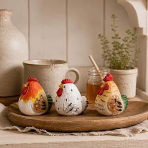 May include: Three small, hand-carved wooden chicken figurines arranged on a wooden tray. The left chicken is orange and white, the centre one is white with black markings, and the right one is tan with brown details. Each has a red comb and wattle.
