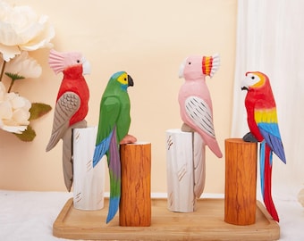 Puede incluir: Cuatro figuras de loros de madera coloridas. Los loros están posados sobre soportes cilíndricos de madera y blancos, dispuestos en una bandeja de madera. Los loros presentan plumaje rojo, verde, rosa y amarillo con tallado detallado. El fondo es beige suave con flores blancas.