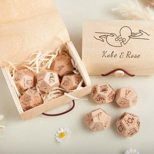 Può includere: Una scatola di legno con coperchio, contenente diversi dadi di legno con immagini e testo incisi. Il coperchio è inciso con un disegno a puzzle a forma di cuore e i nomi "Kobe & Rose". I dadi sono sparsi.