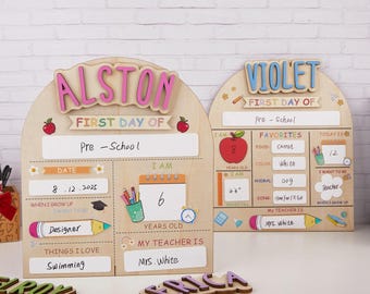 Könnte beinhalten: Holzschilder für den ersten Schultag für Alston und Violet. Die Schilder haben farbenfrohe Texte und Illustrationen, darunter Äpfel, Bleistifte und ein Kalender. Das Alston-Schild enthält den Text "Designer" und "Swimming".