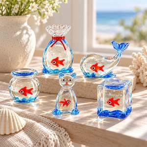 Puede incluir: Una colección de cinco figuras de vidrio transparente con motivos de peces dorados en su interior. El conjunto incluye un cuenco redondo, una bolsa, una ballena, un gato y un cubo, todos con bases de vidrio azul y burbujas de aire.