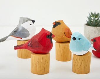 Puede incluir: Cinco figuritas de pájaros de madera, cada una pintada de un color diferente, se muestran en pequeñas bases de madera. Los pájaros son un carbonero gris y blanco, un cardenal rojo, un cardenal amarillo y naranja, un pájaro azul y un pájaro rojo.