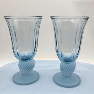 May include: Two light blue stemmed glasses. The glasses have a fluted design on the upper portion and a spiral design on the stem. The base is a solid, round, frosted light blue. The glasses are on a white surface.