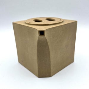 May include: A tan-colored, cube-shaped ceramic container with a curved front and a flat top. The top features two circular openings. A small square opening is visible on the front. The container has a textured surface.
