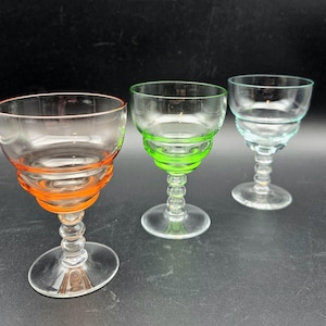 May include: Three clear glass stemmed cocktail glasses with colorful accents. One glass has orange detailing, another green, and the third has blue. The glasses have a round bowl and a decorative stem.