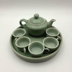 May include: A celadon green ceramic tea set. The set includes a teapot with a lid, six teacups, and a round serving tray. The tea set is a soft, muted green color and is arranged on a white background.
