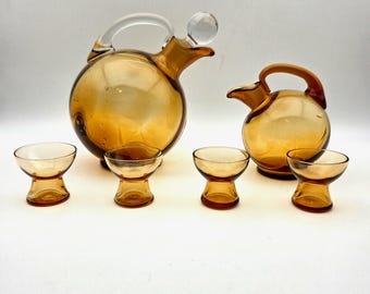 May include: A vintage amber glass drinkware set. The set includes two spherical pitchers with handles and spouts, and five small, stemmed glasses. The glass has a warm, golden hue, and the design is simple and elegant.