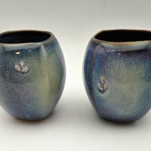 May include: Two ceramic vases with a unique, organic shape. The vases have a blend of blue, green, and purple hues, with a dark brown interior. Each vase features a small, textured design on the front, adding a touch of detail.