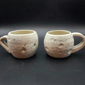 May include: Two ceramic mugs with a textured, off-white surface, resembling the moon's surface with craters. Each mug has a rounded body and a curved handle. The mugs are displayed against a dark background.