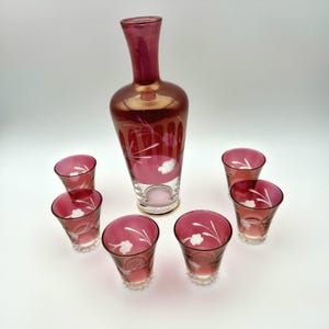 May include: A vintage cranberry glass decanter set. The set includes a tall decanter and six matching glasses. The decanter and glasses feature white floral designs. The set is displayed on a white surface.