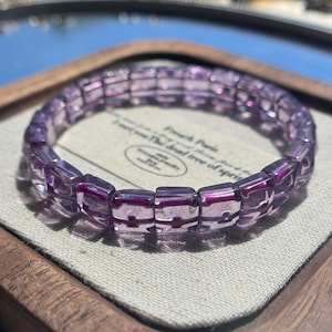 May include: A circular bracelet composed of translucent, light purple square beads. Each bead features a distinct, dark magenta cross-shaped inlay at its center. The beads are strung together in a continuous loop, creating a uniform, geometric pattern.