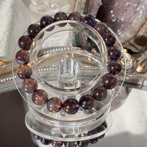 May include: A bracelet with dark purple and brown gemstone beads. The beads are spherical and polished. The bracelet is displayed on a clear acrylic stand, reflecting on a mirrored surface.