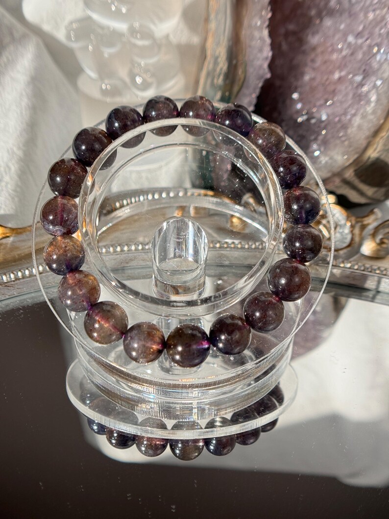 May include: A bracelet with dark purple and brown gemstone beads. The beads are spherical and polished. The bracelet is displayed on a clear acrylic stand, reflecting on a mirrored surface.