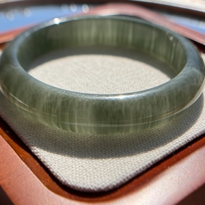 May include: A translucent green jade bangle bracelet. The bracelet has a smooth, polished surface with subtle variations in color and texture. The jade is a soft green hue, with a slightly marbled appearance. The bracelet is circular and appears to be a solid piece of jade.