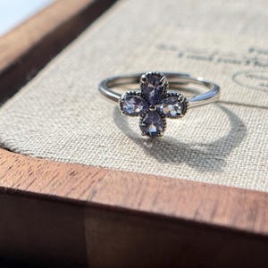 May include: A silver ring featuring a four-petal flower design. Each petal is composed of a light purple gemstone. The ring is set on a neutral-colored surface, with a wooden box in the background.