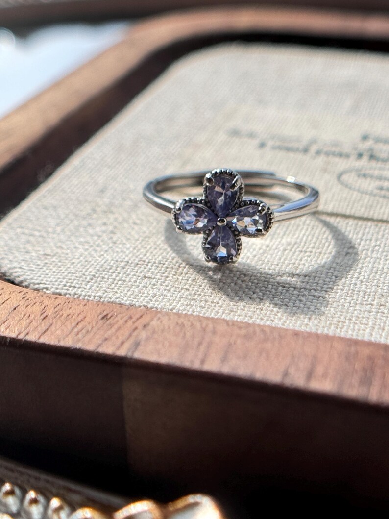 May include: A silver ring featuring a four-petal flower design. Each petal is composed of a light purple gemstone. The ring is set on a neutral-colored surface, with a wooden box in the background.