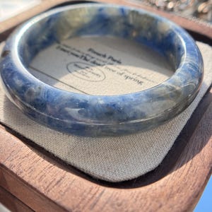 May include: A blue and white marbled bangle bracelet. The bracelet is a solid, circular band with a polished finish. The bracelet is resting on a linen surface inside a wooden tray.