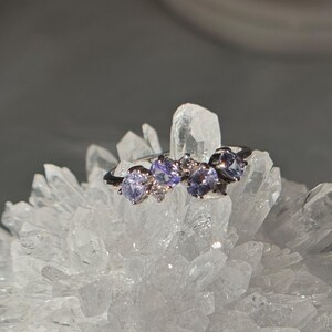 May include: A silver ring featuring three oval, light purple gemstones and small clear accent stones. The ring is set against a backdrop of a cluster of clear quartz crystals, with a soft gray background.