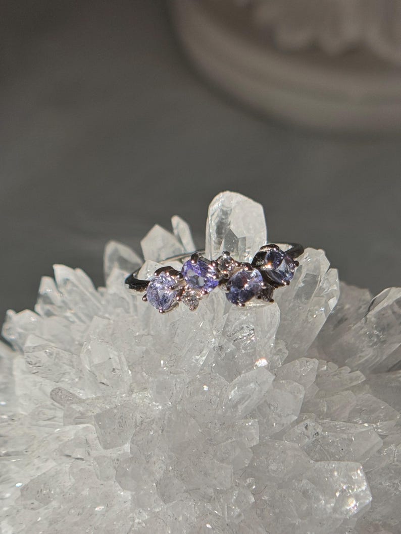 May include: A silver ring featuring three oval, light purple gemstones and small clear accent stones. The ring is set against a backdrop of a cluster of clear quartz crystals, with a soft gray background.