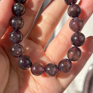 May include: A bracelet made of dark purple, spherical gemstones. The beads have a translucent appearance, with flecks of lighter purple and brown. The bracelet is strung, forming a flexible accessory. The bracelet is held in a hand.