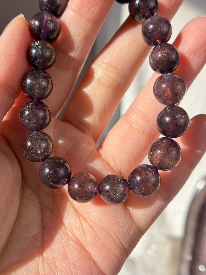 May include: A bracelet made of dark purple, spherical gemstones. The beads have a translucent appearance, with flecks of lighter purple and brown. The bracelet is strung, forming a flexible accessory. The bracelet is held in a hand.