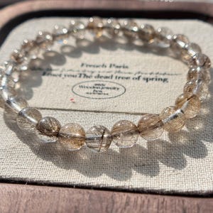 May include: A clear quartz bracelet with gold inclusions. The bracelet is made of round beads and is displayed in a small wooden box with a white fabric lining. The box has a label that reads "French Paris Find you The deal tree of spring."