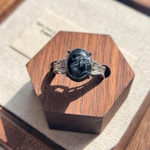 May include: A silver ring featuring an oval-shaped, dark blue and black patterned stone set in a four-prong mount. The band displays a textured, intricate metalwork design that extends from the setting along the sides of the ring.