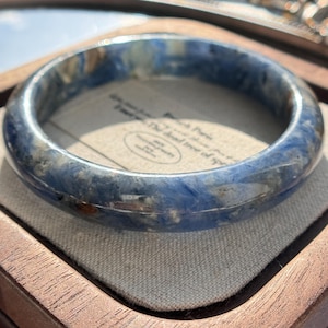 Puede incluir: Un brazalete de piedra preciosa veteada azul y blanca. El brazalete circular tiene un acabado pulido y brillante. Los colores son una mezcla de azul profundo y blanco, creando una apariencia natural similar a la piedra. El brazalete descansa sobre una tela de color neutro.