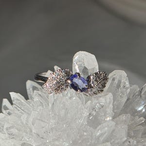 May include: A silver ring with a marquise-cut, deep blue gemstone at the center. The ring features leaf-like designs on either side, embellished with small, clear stones. The ring is displayed on a cluster of clear crystals, creating a sparkling effect.