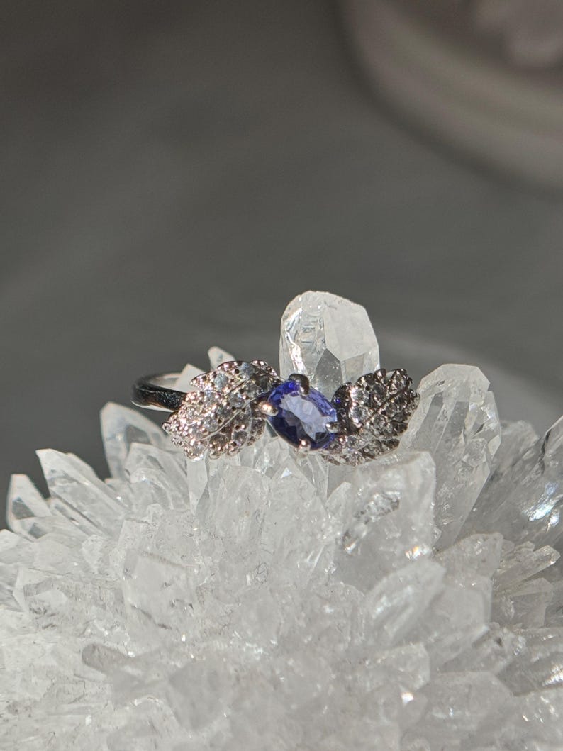 May include: A silver ring with a marquise-cut, deep blue gemstone at the center. The ring features leaf-like designs on either side, embellished with small, clear stones. The ring is displayed on a cluster of clear crystals, creating a sparkling effect.