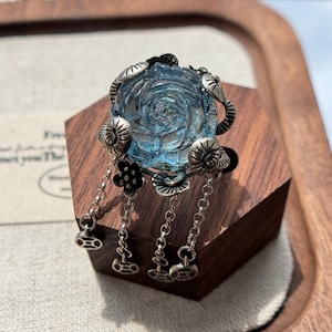 May include: A silver-toned ring featuring a central translucent light blue carved rose gemstone. The setting is adorned with small silver floral accents and four dangling silver chains, each ending in a small circular charm with a square cutout.