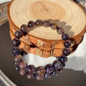 May include: A bracelet made of dark purple and brown gemstone beads. The beads are displayed on a wooden slice, reflecting on a mirrored surface. The beads have a polished, translucent appearance.