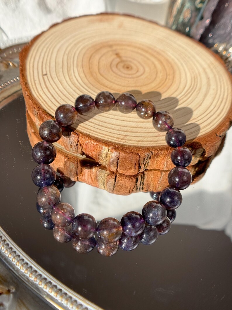 May include: A bracelet made of dark purple and brown gemstone beads. The beads are displayed on a wooden slice, reflecting on a mirrored surface. The beads have a polished, translucent appearance.