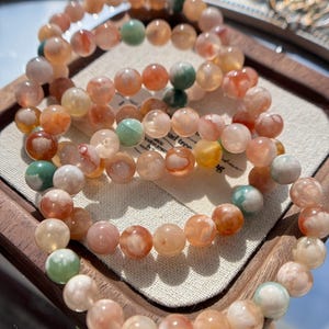Puede incluir: Varios brazaletes con cuentas redondas y coloridas en tonos naranja, melocotón, verde y blanco. Los brazaletes se exhiben en una bandeja de madera con un forro de tela clara. Las cuentas parecen estar hechas de un material translúcido.